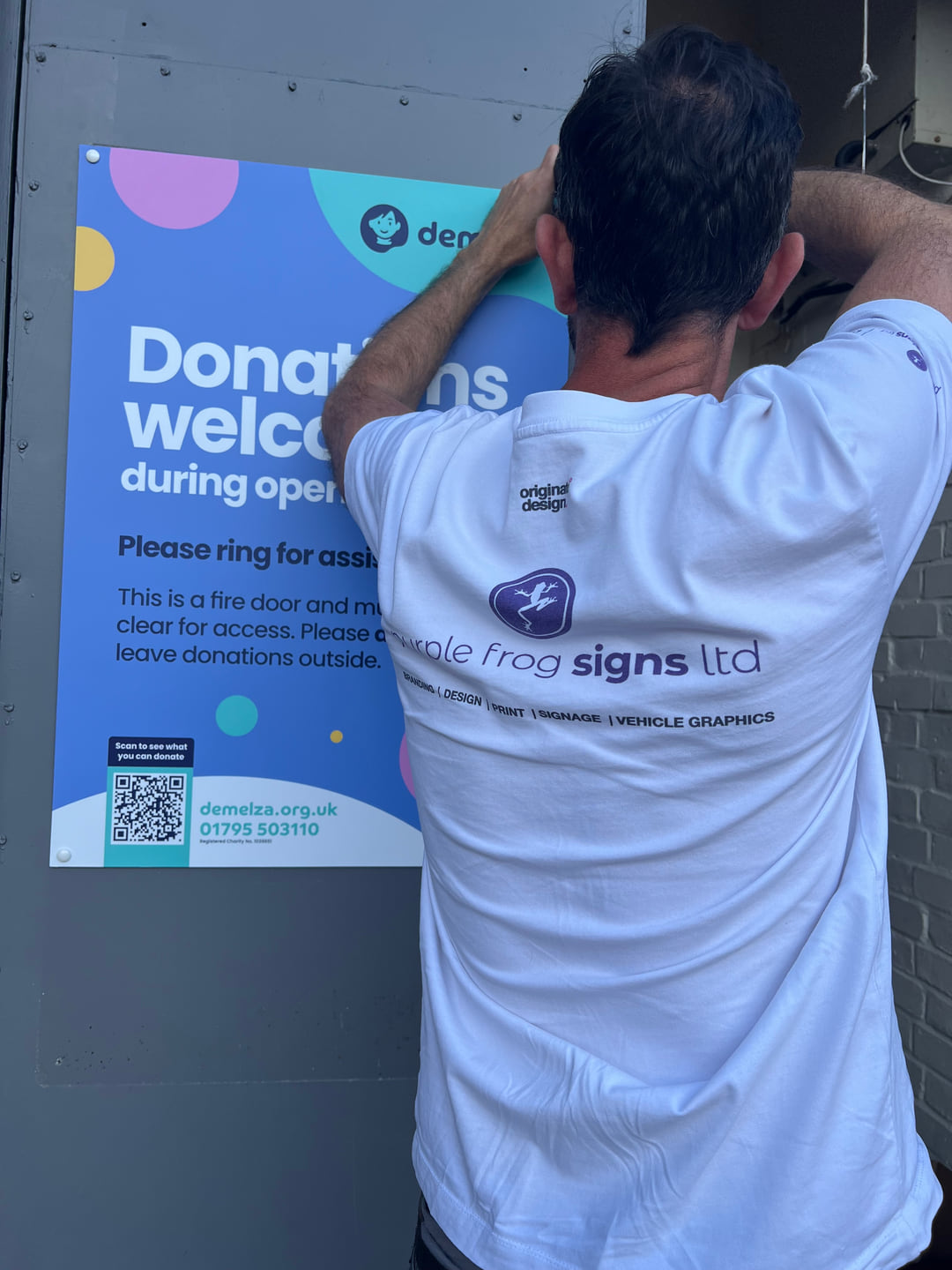 A Purple Frog Signs installer fitting a blue Demelza donation sign to an exterior panel