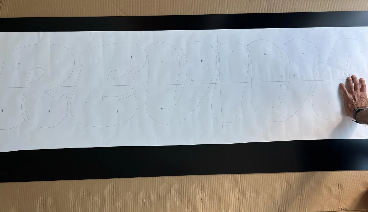 Full-size paper template showing the outlined REVIVAL lettering and stand-off fixing positions on a matt black ACM fascia panel during manufacture of the Thrift & Thrive shopfront sign.