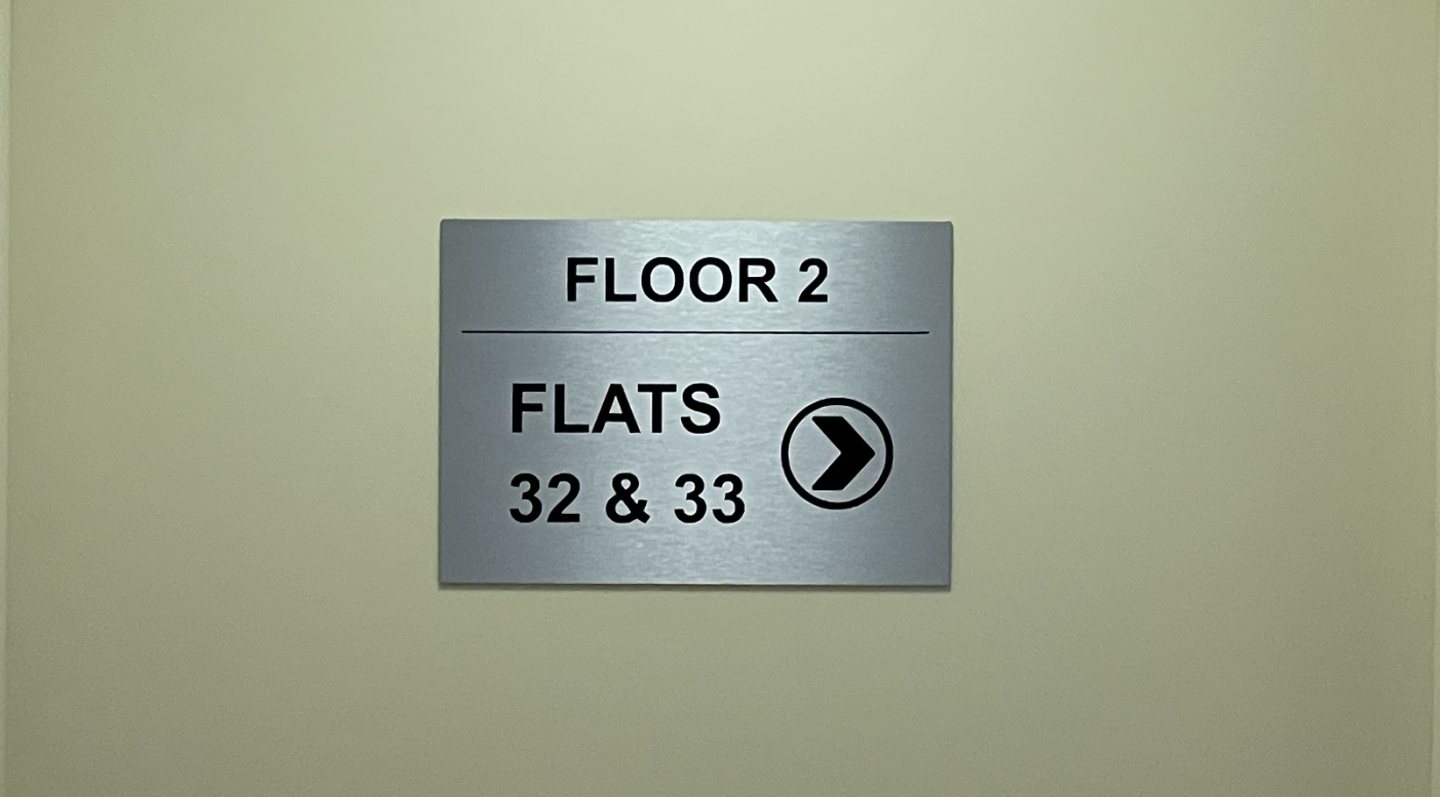 Brushed aluminium composite wayfinding sign with black vinyl lettering and directional arrow for Flats 32 and 33 on Floor 2.