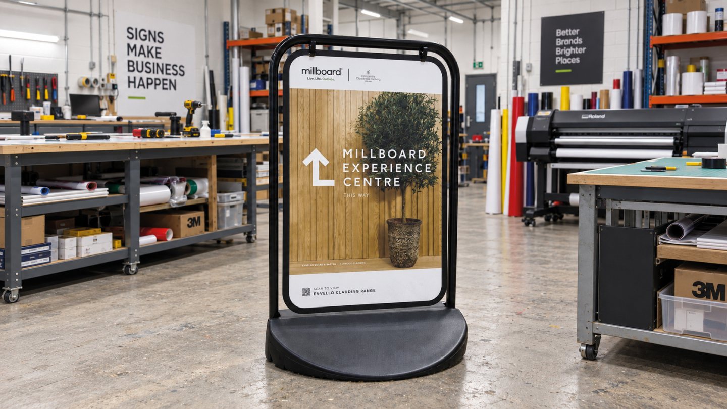 Millboard pavement sign with printed directional graphics, displayed inside a professional signage workshop with workbenches, vinyl rolls and large-format printing equipment.