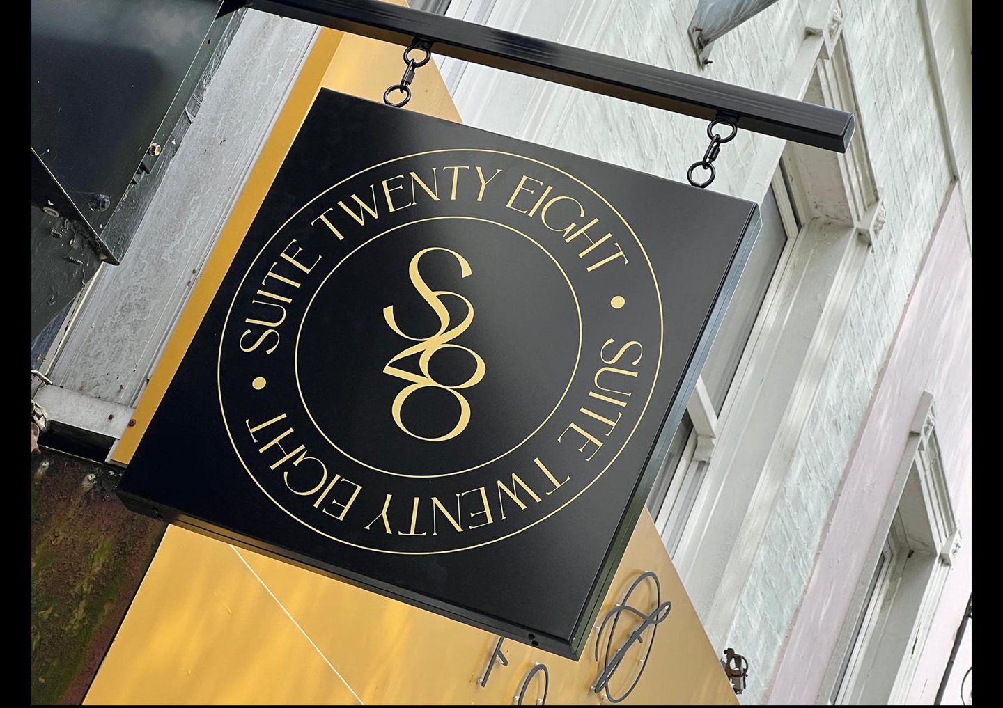 Black projecting shop sign with gold Suite Twenty Eight branding, suspended from a black metal bracket outside a retail premises.